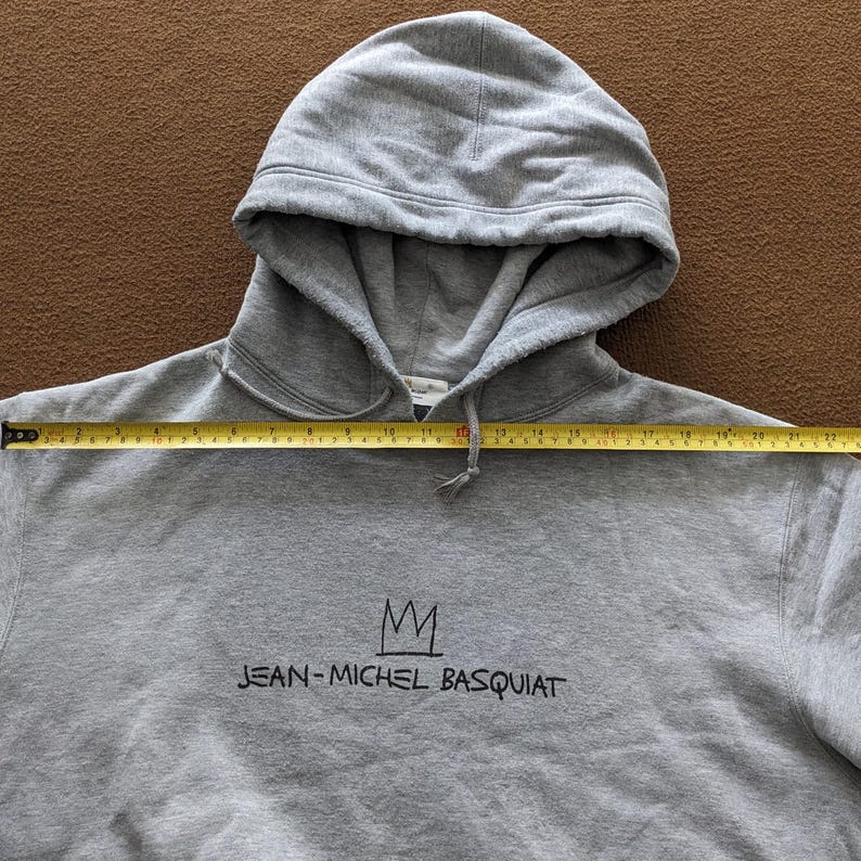Puede incluir: Sudadera con capucha gris con una corona negra y el texto "JEAN-MICHEL BASQUIAT" impreso en la parte delantera.