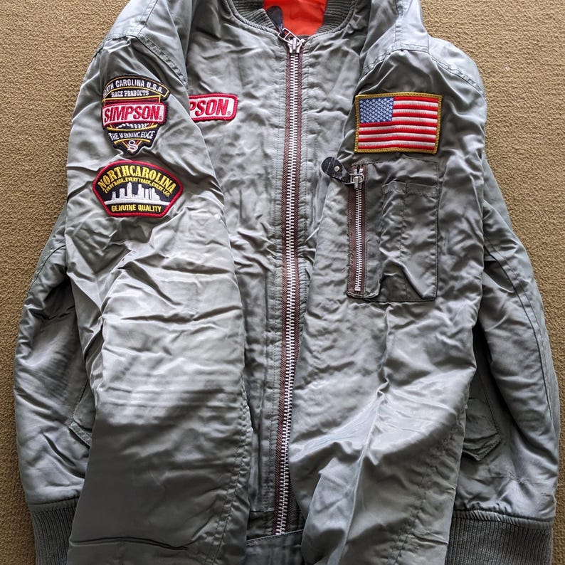 May include: A grey bomber jacket with a zip fastening and several patches. The patches include a North Carolina skyline, a Simpsons logo, and a US flag.