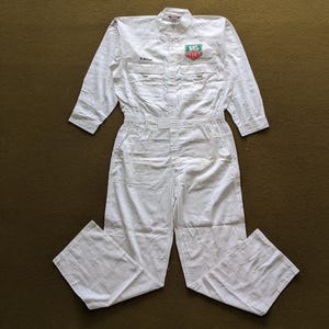 Puede incluir: Mono blanco de algodón con mangas largas y cierre de cremallera. El mono tiene un parche en el pecho con el texto "TAG HEUER" en verde y rojo.