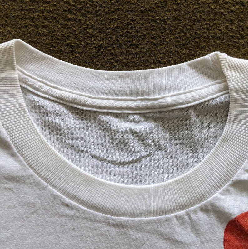 May include: Close-up of the neckline of a white t-shirt. The neckline is a crew neck and has a ribbed texture.
