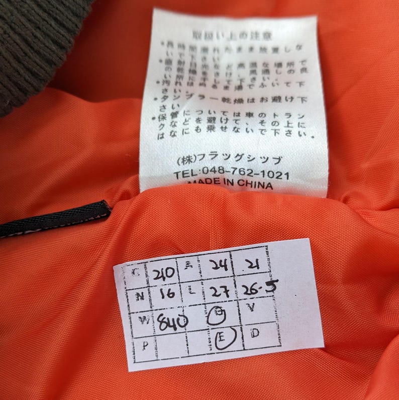 May include: Close-up of a white tag with black text that reads "(株)フラッグシップ TEL:048-762-1021 MADE IN CHINA". Below the tag is a white tag with black text and numbers. The tag reads "C 210 A 24 21 N 16 L 27 26-5 W 840 E V D P".