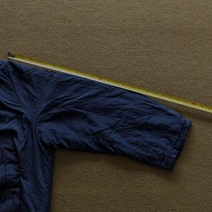 Puede incluir: Una manga de chaqueta azul marino se mide con una cinta m&eacute;trica amarilla. La chaqueta est&aacute; hecha de un material ligero, posiblemente nailon. La manga es parcialmente visible, mostrando el pu&ntilde;o y parte del hombro.