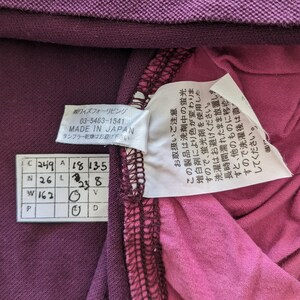 May include: Close-up of a purple garment with attached tags. One tag reads "MADE IN JAPAN" and includes Japanese text. Another tag provides care instructions. The garment's fabric is a deep purple and pink.