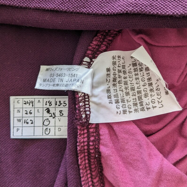 May include: Close-up of a purple garment with attached tags. One tag reads "MADE IN JAPAN" and includes Japanese text. Another tag provides care instructions. The garment's fabric is a deep purple and pink.