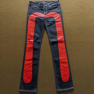 Puede incluir: Vaqueros de mezclilla azul oscuro con un diseño rojo curvo en cada pierna.
