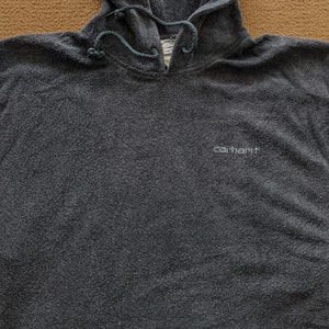 Op de afbeelding: Een donkergrijze hoodie met het woord "carhartt" in wit geborduurd op de voorkant.