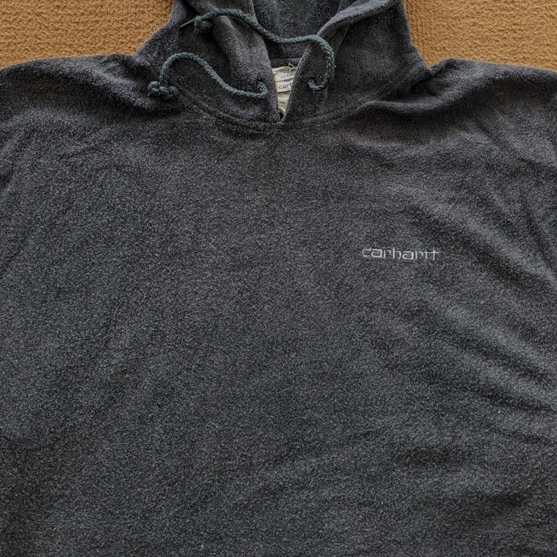 Op de afbeelding: Een donkergrijze hoodie met het woord "carhartt" in wit geborduurd op de voorkant.