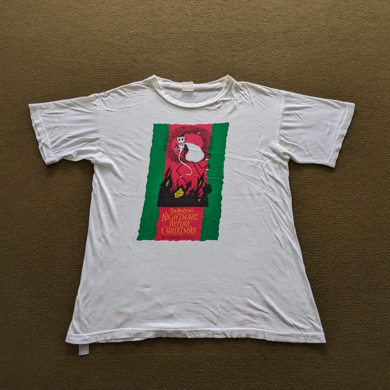 K&ouml;nnte beinhalten: Wei&szlig;es T-Shirt mit einem gr&uuml;nen und roten Aufdruck von Jack Skellington aus dem Film "Nightmare Before Christmas". Der Aufdruck zeigt den Filmtitel und eine Silhouette von Jack Skellington, der eine Tasche h&auml;lt.