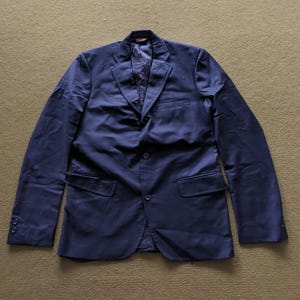 Puede incluir: Un blazer azul marino con cierre de dos botones, solapas con muescas y dos bolsillos delanteros. La chaqueta tiene un corte entallado y mangas largas con pu&ntilde;os abotonados. El blazer est&aacute; hecho de una tela lisa.