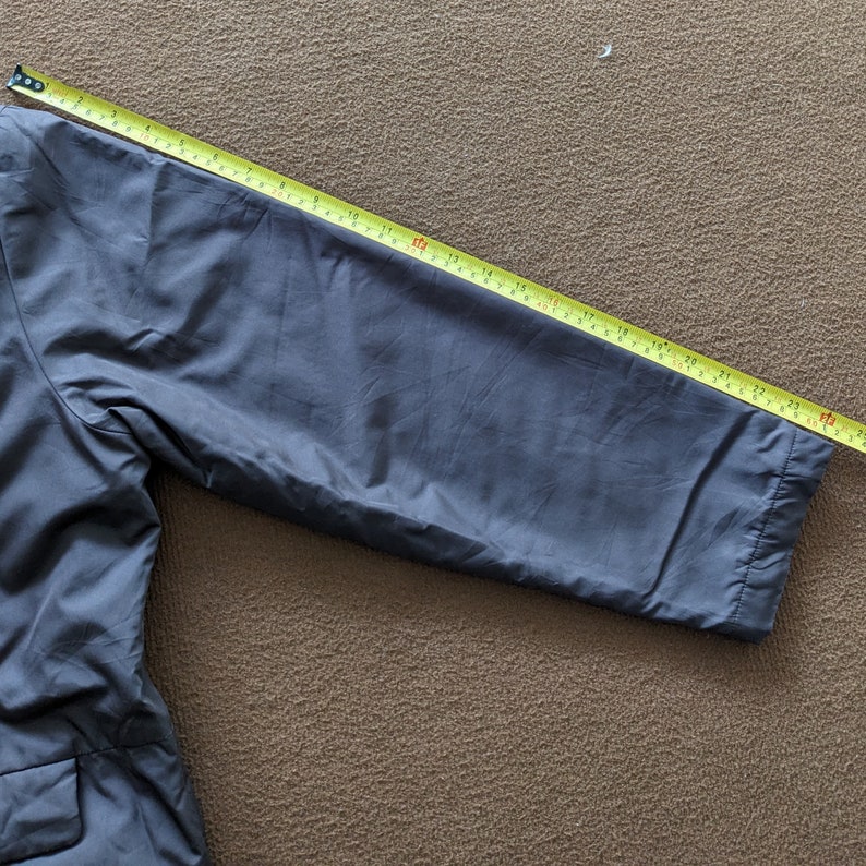 May include: A dark gray jacket with a long sleeve. The sleeve is measured with a yellow tape measure, showing a length of 25 inches.