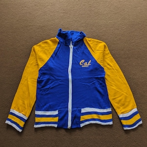 Könnte beinhalten: Eine blaue und gelbe Jacke mit Reißverschluss und weißen Streifen. Auf der Vorderseite der Jacke ist "Cal Berkeley" gestickt.