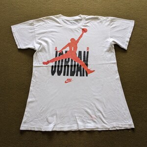 May include: White t-shirt with a red and black graphic of a basketball player with the word "JORDAN" printed in black letters and "NIKE" printed in black letters below the graphic.