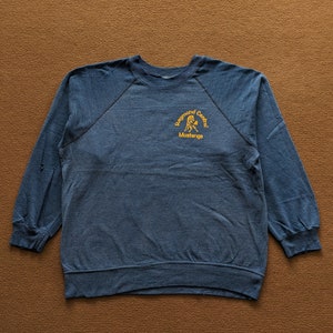 Puede incluir: Una sudadera azul marino con cuello redondo y mangas largas. El texto "Raymond Central Mustangs" está impreso en amarillo en la parte superior izquierda del pecho. La sudadera está hecha de un material suave y cómodo.