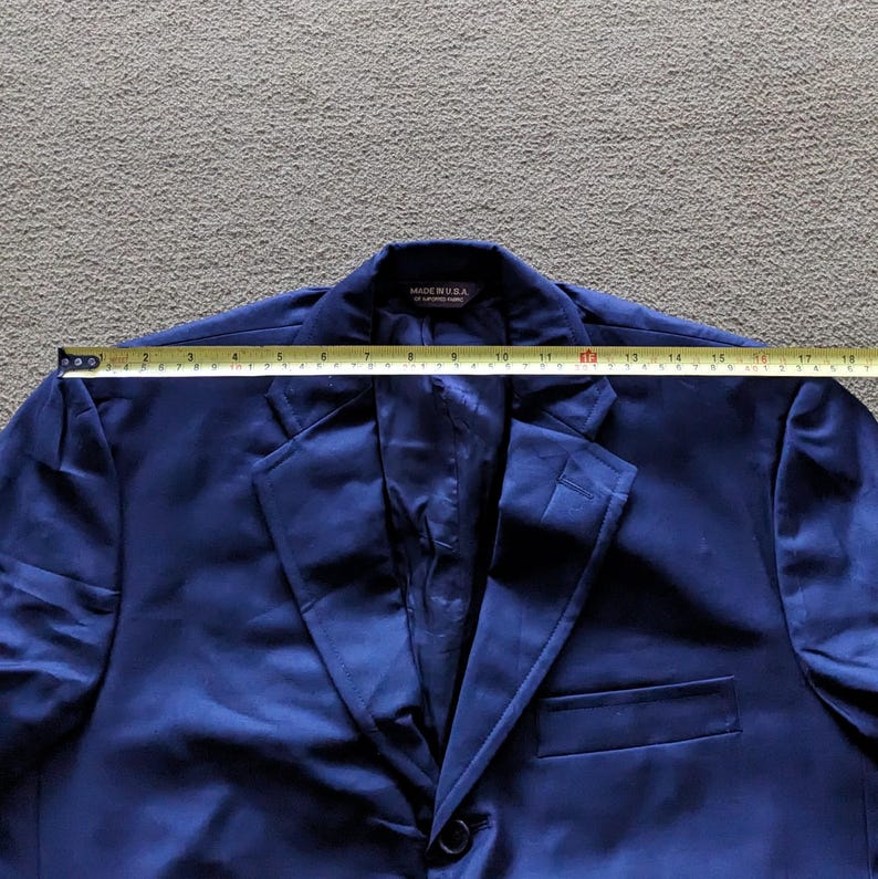 Puede incluir: Un blazer azul marino con cierre de un solo bot&oacute;n y solapa con muescas. El blazer se mide con una cinta m&eacute;trica. La etiqueta dice "MADE IN USA". El ancho total del blazer es de aproximadamente 45,7 cm.