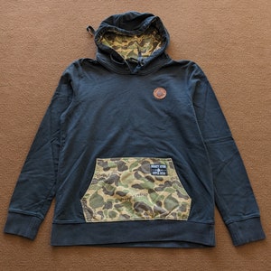 May include: A dark blue hooded sweatshirt with a camouflage pocket and a small brown patch on the chest. The pocket has a small patch with the text "Mickey Atom Super Hero" on it.