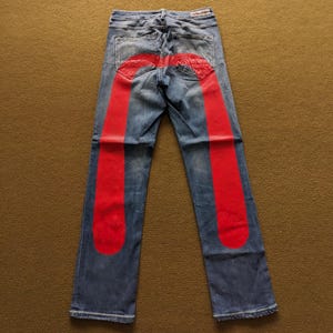 Puede incluir: Vaqueros de mezclilla azul con un diseño gráfico rojo en las piernas. El diseño gráfico es una forma grande y curva que se asemeja a la letra U.