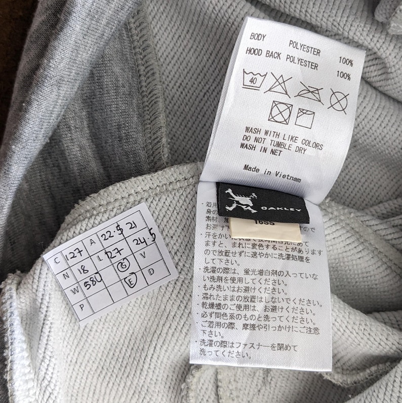 以下が含まれることがあります： 白い洗濯表示タグと白いサイズタグが付いたグレーのフード付きスウェットシャツ。洗濯表示タグには、衣類の洗濯、乾燥、アイロンがけに関する指示が記載されています。サイズタグには、スウェットシャツのサイズと寸法が記載されています。タグには、「Oakley」と「1055」の文字も記載されています。