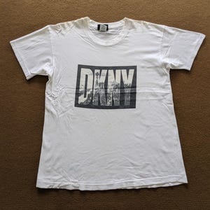 May include: White t-shirt with a rectangular graphic displaying the letters "DKNY" in a bold font. The graphic incorporates a black and white image of a city skyline. The t-shirt has short sleeves and a crew neck.