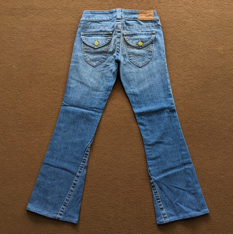 May include: A pair of blue denim jeans with a bootcut leg. The jeans have a brown leather patch on the back with the text "True Religion" in white. The jeans have yellow stitching and two small pockets on the back.