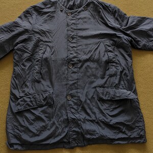 Puede incluir: Chaqueta azul marino oscuro con cierre de botones, dos bolsillos delanteros y cuello mandar&iacute;n. La chaqueta tiene una textura ligeramente arrugada y un ajuste relajado. La prenda est&aacute; tendida sobre una superficie color mostaza.