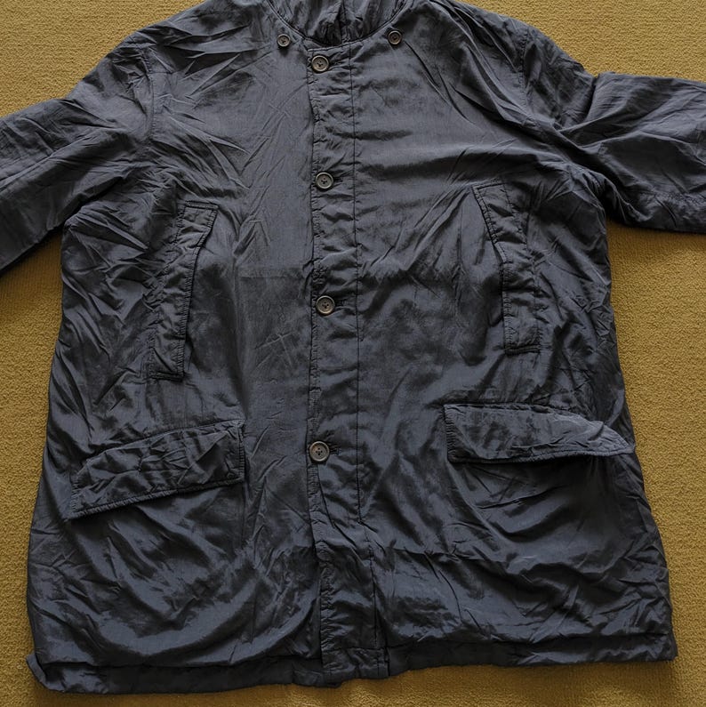Puede incluir: Chaqueta azul marino oscuro con cierre de botones, dos bolsillos delanteros y cuello mandar&iacute;n. La chaqueta tiene una textura ligeramente arrugada y un ajuste relajado. La prenda est&aacute; tendida sobre una superficie color mostaza.