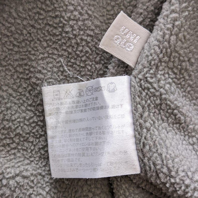 Puede incluir: Una etiqueta de cuidado blanca con texto japon&eacute;s y s&iacute;mbolos para las instrucciones de lavado. La etiqueta est&aacute; unida a una tela de polar gris.