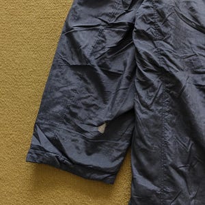 Puede incluir: Primer plano de una secci&oacute;n de una chaqueta azul oscuro. La tela tiene una textura brillante y ligeramente arrugada. Son visibles costuras y una peque&ntilde;a zona de desgaste. El fondo es una superficie texturizada de color marr&oacute;n dorado.