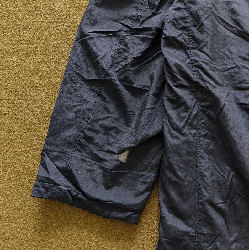 Puede incluir: Primer plano de una secci&oacute;n de una chaqueta azul oscuro. La tela tiene una textura brillante y ligeramente arrugada. Son visibles costuras y una peque&ntilde;a zona de desgaste. El fondo es una superficie texturizada de color marr&oacute;n dorado.