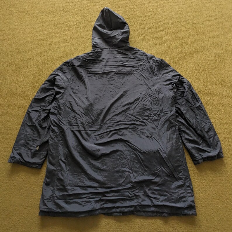 Puede incluir: Chaqueta con capucha gris oscuro, vista desde atr&aacute;s, sobre una superficie amarillo-verdosa. La chaqueta tiene mangas largas y una capucha. La tela parece ligera y ligeramente arrugada.