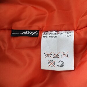 May include: A close-up of the inside of a bright orange jacket with a black label that reads "Approved by NORIK" and a white label with Japanese text and care instructions. The care instructions include a 30 degree Celsius washing symbol, a dry clean symbol, and an iron symbol.