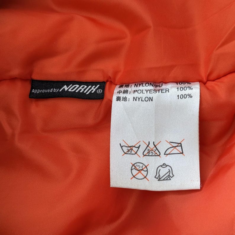 May include: A close-up of the inside of a bright orange jacket with a black label that reads "Approved by NORIK" and a white label with Japanese text and care instructions. The care instructions include a 30 degree Celsius washing symbol, a dry clean symbol, and an iron symbol.