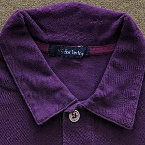 May include: Close-up of a purple polo shirt with a collared neckline and a two-button placket. The shirt features the text "Y's for living" on a small tag at the back of the neck. The fabric appears to be a textured knit.