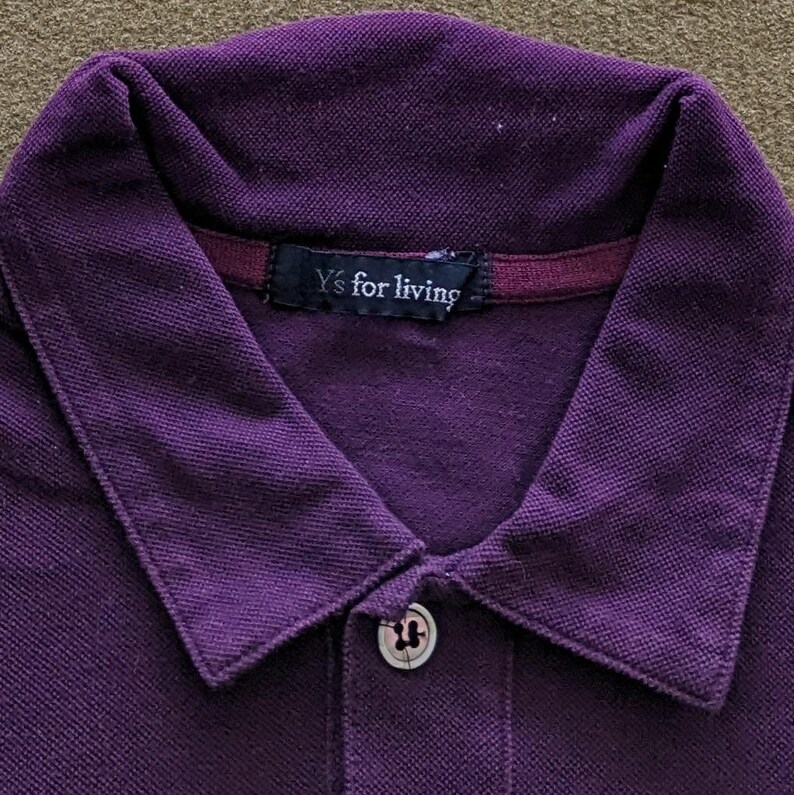 May include: Close-up of a purple polo shirt with a collared neckline and a two-button placket. The shirt features the text "Y's for living" on a small tag at the back of the neck. The fabric appears to be a textured knit.