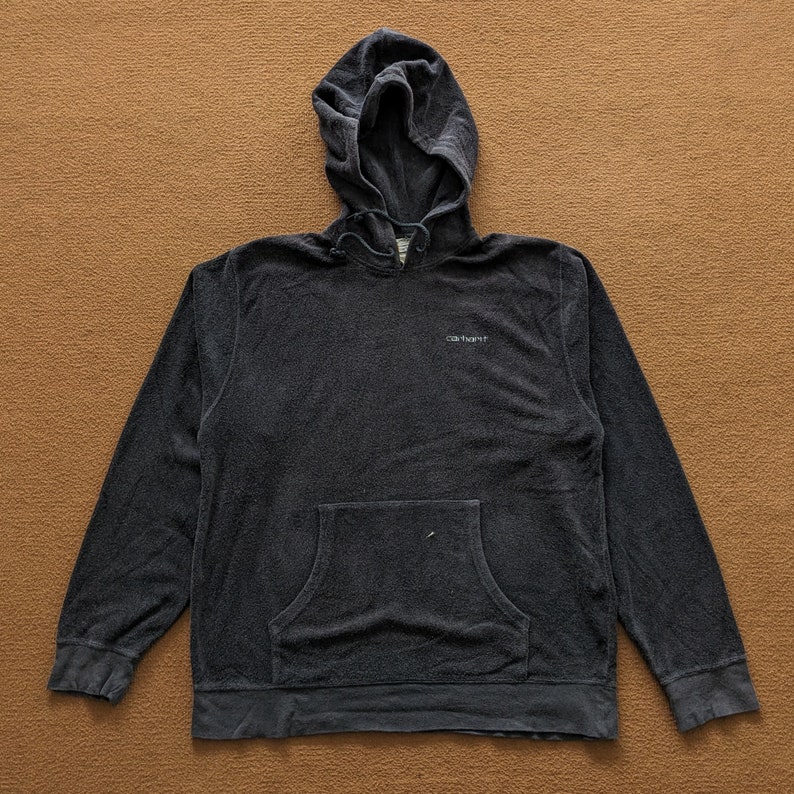 Op de afbeelding: Een donkergrijze hoodie met een voorzak en het woord "Carhartt" geborduurd op de borst.