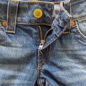 May include: Close-up of a pair of blue denim jeans with a zipper and a yellow button that reads "True Religion Brand Jeans".