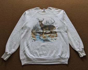 May include: A gray sweatshirt with a graphic print of a white-tailed deer standing in a snowy forest. The deer is brown with a white tail and antlers.