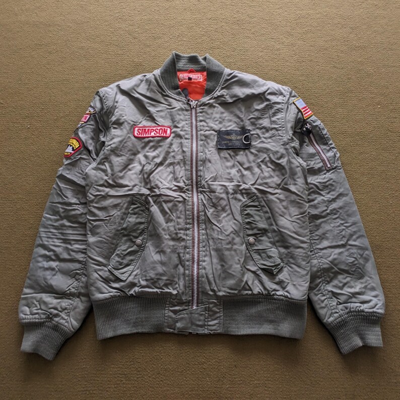 May include: A green bomber jacket with a zip fastening and multiple patches, including a red patch with the word "SIMPSON" on it.