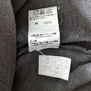 Op de afbeelding: Een zwart katoenen T-shirt met een wit label eraan bevestigd. Het label heeft de tekst "綿 100%" en "企画生産(株)エドウイン商事 03-5604-8900". Er is ook een apart wit label met de tekst "C 87 N 12 W 570 P A 22 19 27 27 V D".