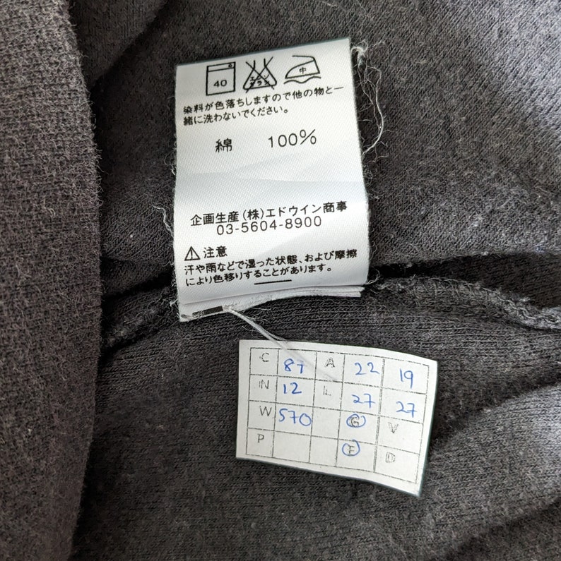 Op de afbeelding: Een zwart katoenen T-shirt met een wit label eraan bevestigd. Het label heeft de tekst "綿 100%" en "企画生産(株)エドウイン商事 03-5604-8900". Er is ook een apart wit label met de tekst "C 87 N 12 W 570 P A 22 19 27 27 V D".