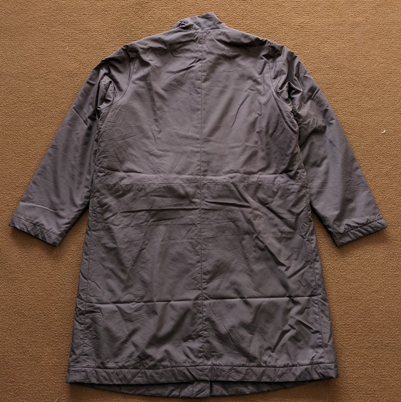 May include: A long, dark gray coat with a stand-up collar and a long zipper. The coat has a relaxed fit and is made of a lightweight fabric.