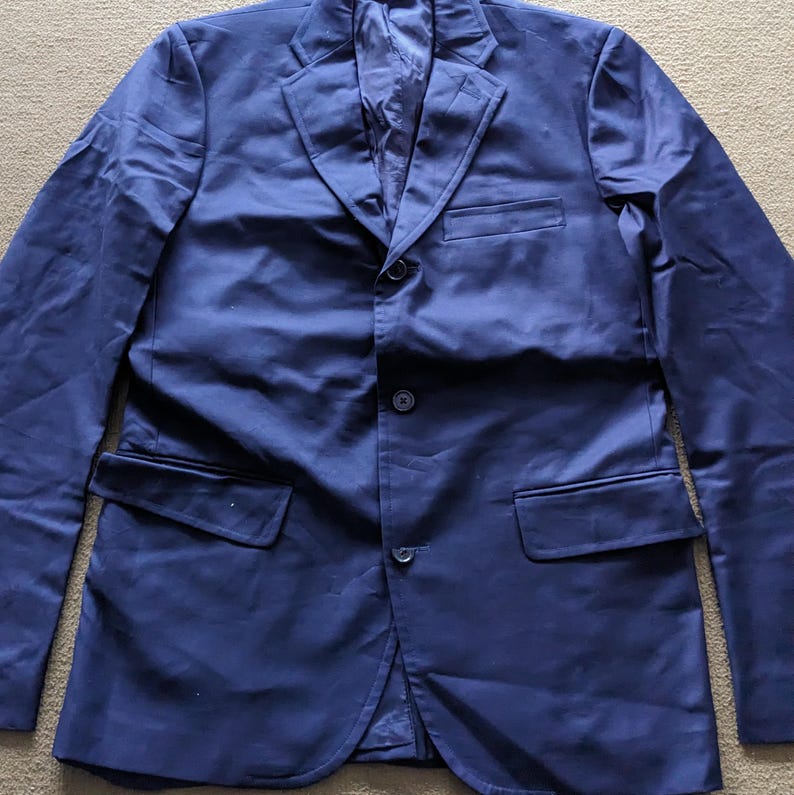 Puede incluir: Un blazer azul marino con cierre de tres botones, solapa de muesca y dos bolsillos delanteros. El blazer est&aacute; hecho de una tela lisa y tiene un dise&ntilde;o cl&aacute;sico y entallado. La prenda est&aacute; colocada sobre una superficie neutra.