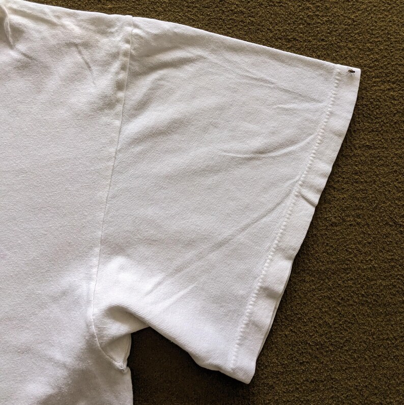 May include: Close-up of the white cotton sleeve of a t-shirt. The sleeve is folded over and the stitching is visible.