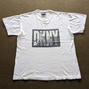 May include: White t-shirt featuring a black and white graphic print of the word "DKNY" with a cityscape in the background.