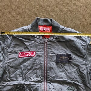 May include: A green bomber jacket with a red "SIMPSON" patch on the left chest and a black patch with "NORTH CAROLINA U.S.A. SINCE 1979" on the right chest. The jacket has a zipper closure and a ribbed collar.