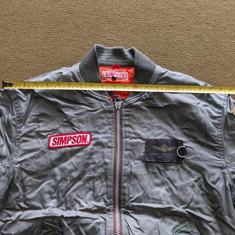 May include: A green bomber jacket with a red "SIMPSON" patch on the left chest and a black patch with "NORTH CAROLINA U.S.A. SINCE 1979" on the right chest. The jacket has a zipper closure and a ribbed collar.