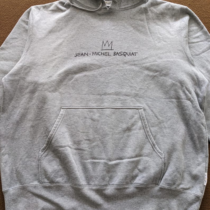 Puede incluir: Una sudadera con capucha gris con una corona negra y el texto "JEAN-MICHEL BASQUIAT" impreso en la parte delantera.