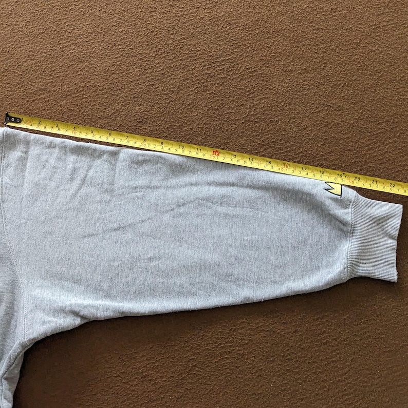 Puede incluir: Una sudadera gris con un logotipo de corona amarilla en la manga. La sudadera est&aacute; colocada sobre una superficie marr&oacute;n con una cinta m&eacute;trica al lado.