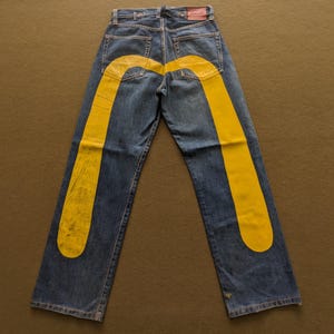 Puede incluir: Vaqueros azules con grandes diseños amarillos curvos en los bolsillos traseros y las piernas. Los vaqueros tienen un diseño clásico de cinco bolsillos y un parche de cuero rojo en la cintura trasera.