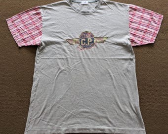 May include: A gray t-shirt with plaid sleeves. The shirt features a circular logo with the text "ITALY C.P. COMPANY" and a wing design. The sleeves are a pink and red plaid pattern.