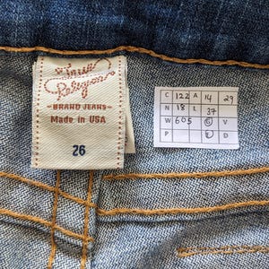 May include: A close-up of a blue denim jean label with the text "True Religion Brand Jeans Made in USA" and the size "26". A white tag with size measurements is also visible.